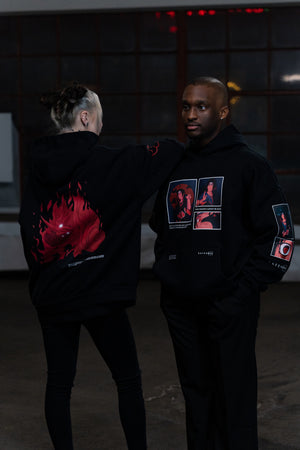 Man and woman in black hoodies with striking red anime art on the back and character patches on the sleeves, posing thoughtfully in a dimly lit urban setting.